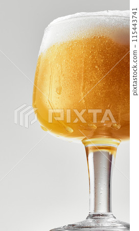 Close-up of mug filled with golden, hazy beer topped with thick, frothy white foam against grey studio background. Close-up of mug filled with golden, hazy beer topped with thick, frothy white foam against grey studio background. 115443741