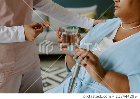 Nurse standing next to African American female patient and offering red pills while elderly woman sitting still and holding cane and glass of water Nurse standing next to African American female patient and offering red pills while elderly woman sitting still and holding cane and glass of water 115444299