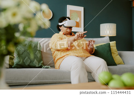 Low angle shot of senior African American woman testing VR goggles while resting on sofa 115444431