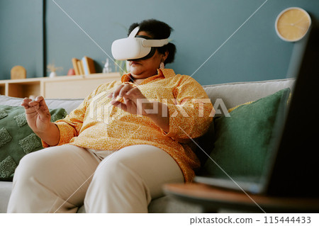 Mature African American woman sitting on sofa and getting in touch with virtual reality 115444433