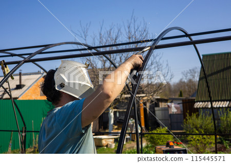 Assembling a frame for a greenhouse by welding in the garden. a man with a protective mask fastens metal pipes with electric arc welding. A man prepares a greenhouse by welding the frame. Assembling a frame for a greenhouse by welding in the garden. a man with a protective mask fastens metal pipes with electric arc welding. A man prepares a greenhouse by welding the frame. 115445571