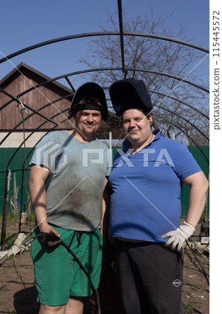 Two cute male welders look at the camera and smile. Men weld a frame for a greenhouse in the garden. Two men in protective welding masks stand in a vegetable garden and look at the camera. 115445572