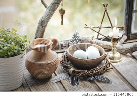 Home atmospheric spring altar with willow wreath, clay jug with milk, bowl with eggs, the first greens in the pot, candle and birds feathers on wooden surface, selected focus. 115445905