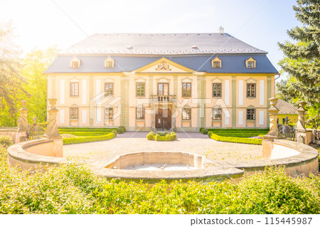 Photo of Potstejn, a charming baroque chateau in Czechia. Yellow facade, dark blue roof, surrounded by lush greenery, trees, and a garden. 115445987
