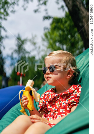 Little girl eating a banana. Portrait of a fashionable, stylish beautiful baby girl in sunglasses and a red dress while sitting in a bean bag chair 115446966