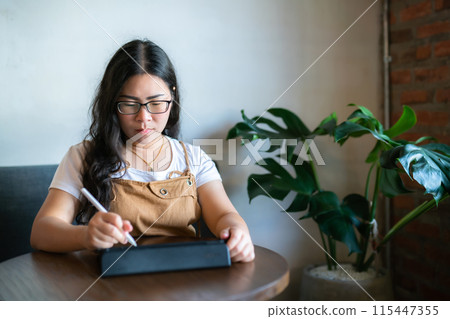 Portrait asian freelance people business female wearing glasses stylish hipste Draw or taking note on digital tablet with electronic pen for browsing internet, chatting and blogging in coffee shop. 115447355