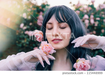 Fairy tale woman portrait. Young beautiful woman Fairy holds pink flower rose on blooming bush background. Hairstyle loose green hair, romantic purple medieval vintage dress. Summer fantasy garden. 115448999