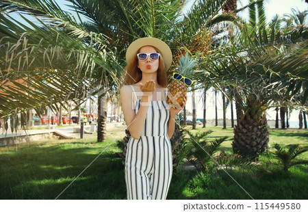 Summer fashion outfit of trendy young woman holding pineapple posing in park with green palm trees 115449580