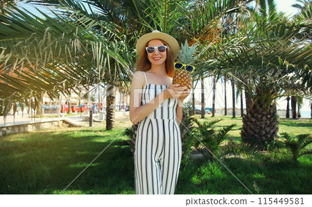 Summer fashion outfit of stylish happy smiling young woman holding pineapple in park with palm trees 115449581