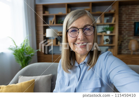 Senior woman with glasses taking a selfie at home in a modern, cozy living room environment. She is smiling and looking directly at the camera. Senior woman with glasses taking a selfie at home in a modern, cozy living room environment. She is smiling and looking directly at the camera. 115450410