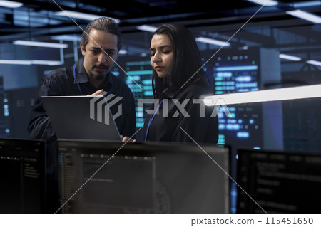 Programmers in server room checking recovery plan, monitoring tech gear energy consumption using notebook. Colleagues in data center workplace doing routine disk checks using laptop and tablet 115451650