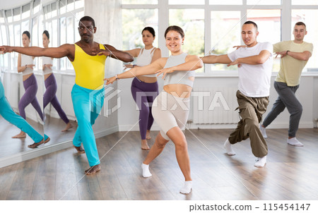 Smiling woman with multinational group active people enthusiastically training social latin cha-cha-cha dance movements in dance salon 115454147