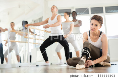 Ballerina ties her pointe shoes on her feet during break between ballet rehearsals. Other participants continue to perform various ballet steps 115454393
