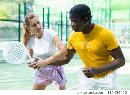 Portrait of caucasian female trainer teaching african american man playing padel 115454420