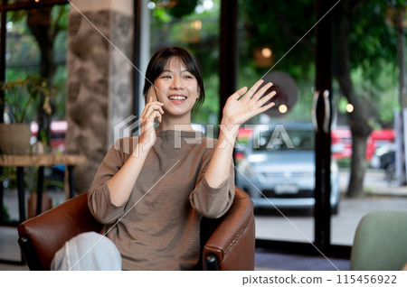 An attractive Asian woman is waving her hand to greet someone while talking on the phone in a cafe. 115456922