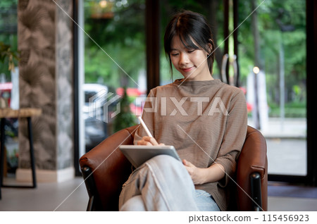 A beautiful Asian woman is writing on her tablet with a stylus pen, working remotely from a cafe. 115456923
