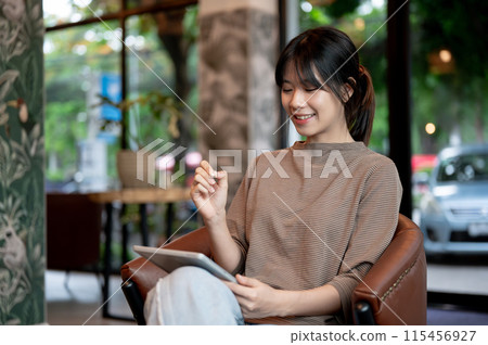 A beautiful Asian woman is holding a stylus pen and working on her digital tablet in a cafe. A beautiful Asian woman is holding a stylus pen and working on her digital tablet in a cafe. 115456927