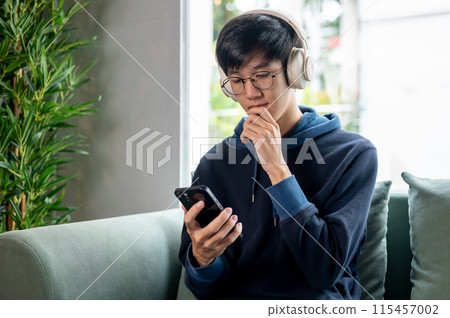 A young, positive Asian man in a hoodie and headphones is using his smartphone on the couch. 115457002