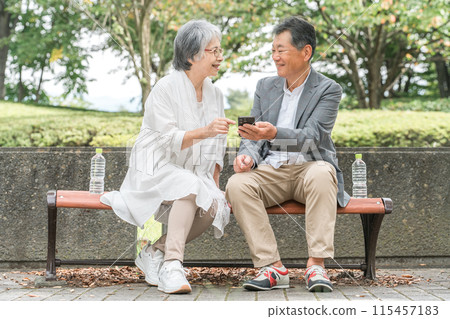 一對老夫婦(老人)在公園的長椅上一邊看智慧型手機一邊聊天 一對老夫婦(老人)在公園的長椅上一邊看智慧型手機一邊聊天 115457183