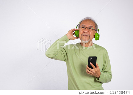 Portrait Asian smiling old man wearing glasses and headphones is holding smartphone listen music in headphones studio shot isolated on white background, enjoying his music Portrait Asian smiling old man wearing glasses and headphones is holding smartphone listen music in headphones studio shot isolated on white background, enjoying his music 115458474