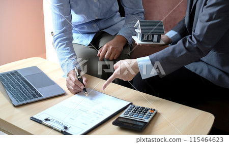 A real estate agent with a house model is talking to clients about buying home insurance and having customers sign contracts under the formal contract agreement. Home rental and insurance concept 115464623