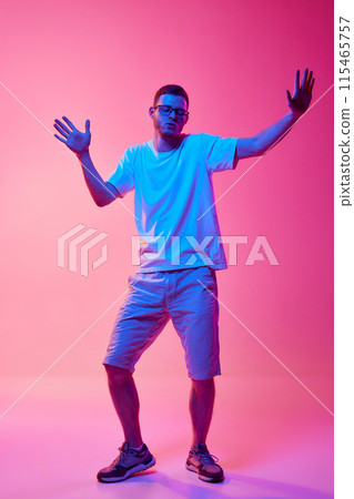 Full-length image of young man in glasses and casual clothes cheerfully dancing and having fun against pink background in neon light 115465757