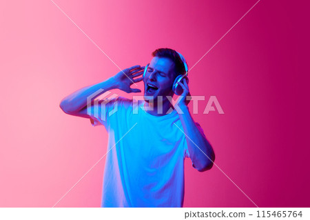 Portrait of young man in white t-shirt listening to music in headphones and singing against pink background in neon light Portrait of young man in white t-shirt listening to music in headphones and singing against pink background in neon light 115465764