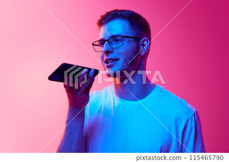 Portrait of young man in glasses and casual clothes using mobile phone for communication, recording voice message against pink background in neon light Portrait of young man in glasses and casual clothes using mobile phone for communication, recording voice message against pink background in neon light 115465790