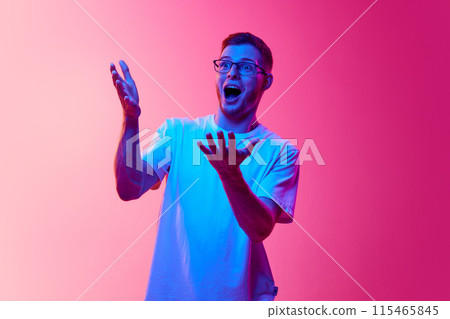 Wow. Young man with glasses, wearing white t-shirt, expressing surprise with his mouth open and hands raised against pink background in neon light Wow. Young man with glasses, wearing white t-shirt, expressing surprise with his mouth open and hands raised against pink background in neon light 115465845