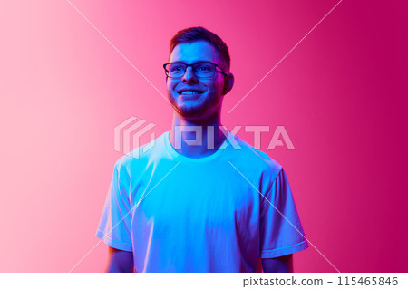 Portrait of young man in casual white t-shirt and glasses posing, looking at camera and smiling against pink background in neon light Portrait of young man in casual white t-shirt and glasses posing, looking at camera and smiling against pink background in neon light 115465846