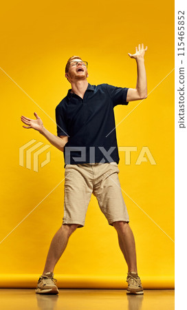 Full-length image of young man in dark blue polo shirt and beige shorts dancing against bright yellow background Full-length image of young man in dark blue polo shirt and beige shorts dancing against bright yellow background 115465852