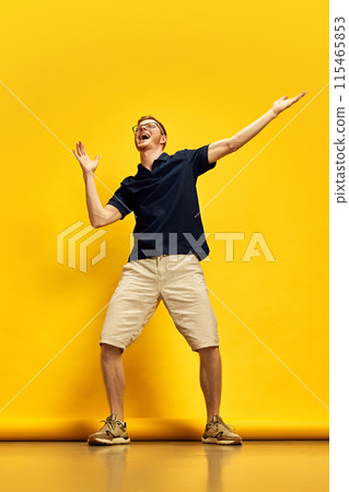 Full-length image of young man in dark blue polo shirt and beige shorts dancing against bright yellow background Full-length image of young man in dark blue polo shirt and beige shorts dancing against bright yellow background 115465853