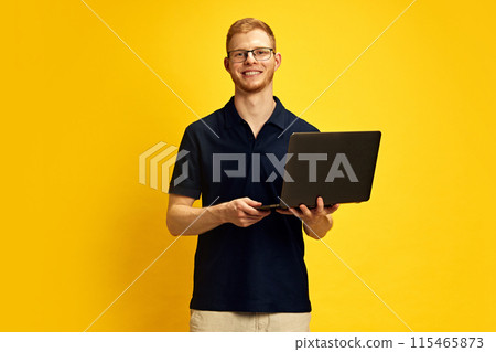 Confident smiling young man in glasses holding laptop against bright yellow background. Business, education and technology. 115465873