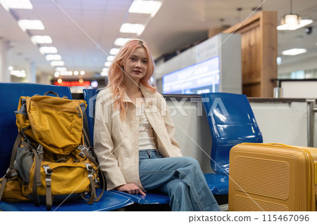 Young Woman Traveling at Airport with Yellow Backpack and Suitcase, Sitting on Blue Chairs in Modern Terminal, Ready for Adventure and Exploration Young Woman Traveling at Airport with Yellow Backpack and Suitcase, Sitting on Blue Chairs in Modern Terminal, Ready for Adventure and Exploration 115467096