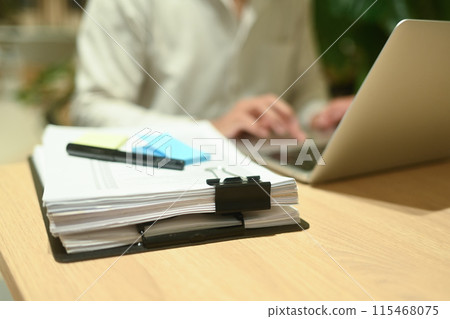 Stack of report paper documents on wooden table with businessman using laptop in background Stack of report paper documents on wooden table with businessman using laptop in background 115468075