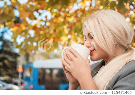 A blonde woman is drinking coffee while sitting under a tree. The scene is peaceful and relaxing, with the woman enjoying her coffee and the natural surroundings. 115471403