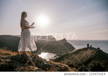 A woman stands on a rocky hill overlooking the ocean. She is wearing a white dress and she is enjoying the view. The scene is serene and peaceful, with the sun shining brightly in the background. A woman stands on a rocky hill overlooking the ocean. She is wearing a white dress and she is enjoying the view. The scene is serene and peaceful, with the sun shining brightly in the background. 115471414