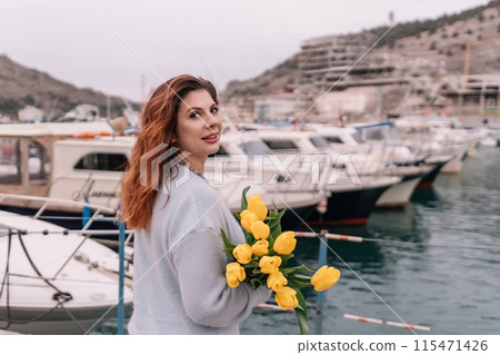 Woman holds yellow tulips in harbor with boats docked in the background., overcast day, yellow sweater, mountains 115471426