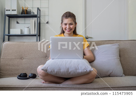 Young Woman Enjoying Social Media on Laptop at Home, Sitting Comfortable on Sofa in Modern Living Room, Smiling and Relaxed 115473905