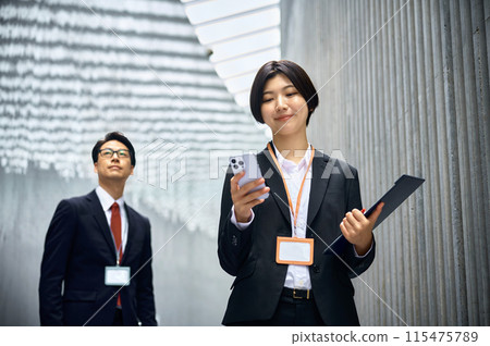 一位身穿西裝的女商人在辦公室走動時操作智慧型手機 一位身穿西裝的女商人在辦公室走動時操作智慧型手機 115475789