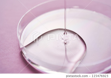 Serum or cosmetic oil flows into a transparent bowl on a purple background. Serum or cosmetic oil flows into a transparent bowl on a purple background. 115476923