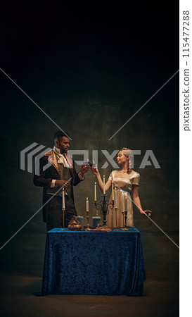 Elegant couple in vintage attire drinking wine over candlelit table against dark, moody background. Romantic evening Elegant couple in vintage attire drinking wine over candlelit table against dark, moody background. Romantic evening 115477288