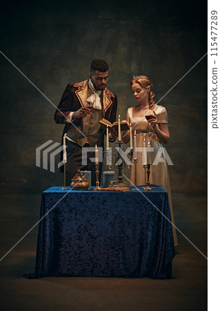 Cozy evening atmosphere. Elegant young woman and handsome man, royal family standing by the table with candles, drinking wine and reading books 115477289