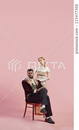 Baroque Elegance. Man in baroque-inspired velvet coat sitting with elegant woman standing by his side against pink studio background 115477308