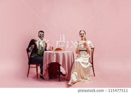 Elegant Victorian couple in period costumes sitting at table and watching movie in 3D glasses, with popcorn against pink studio background Elegant Victorian couple in period costumes sitting at table and watching movie in 3D glasses, with popcorn against pink studio background 115477311