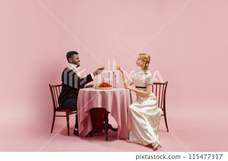 Man and woman, dressed in period attire, share romantic dinner, drinking champagne against pink studio background. 18th-century dining customs Man and woman, dressed in period attire, share romantic dinner, drinking champagne against pink studio background. 18th-century dining customs 115477317