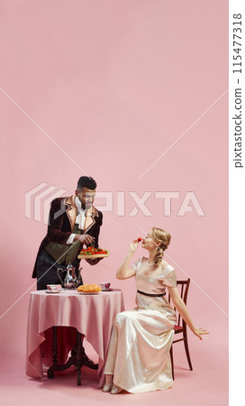 Man in traditional attire presents plate of berries to lady against pink studio background. Gentleman and lady having breakfast. 115477318