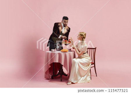 Elegance of the Edwardian Era. Man in period suit serving strawberries to woman against pink studio background. Leisure and luxury of upper-class life. Elegance of the Edwardian Era. Man in period suit serving strawberries to woman against pink studio background. Leisure and luxury of upper-class life. 115477321