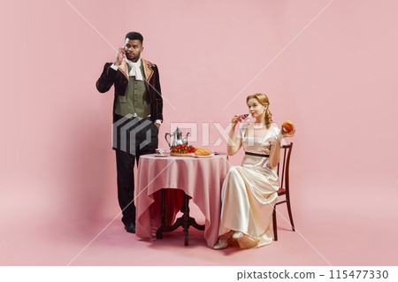 Viscount and Viscountess, man and woman in period costumes against pink studio background. Man talking on mobile phone, woman eating croissant 115477330