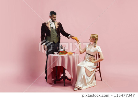 Elegant man and woman in period costumes having breakfast, man giving croissant to attractive woman against pink studio background Elegant man and woman in period costumes having breakfast, man giving croissant to attractive woman against pink studio background 115477347